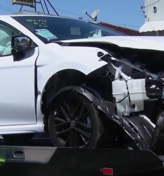 White sedan with severe front-end damage and deployed airbags on a tow truck bed, emergency responder nearby in a neon jacket.