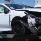White sedan with severe front-end damage and deployed airbags on a tow truck bed, emergency responder nearby in a neon jacket.