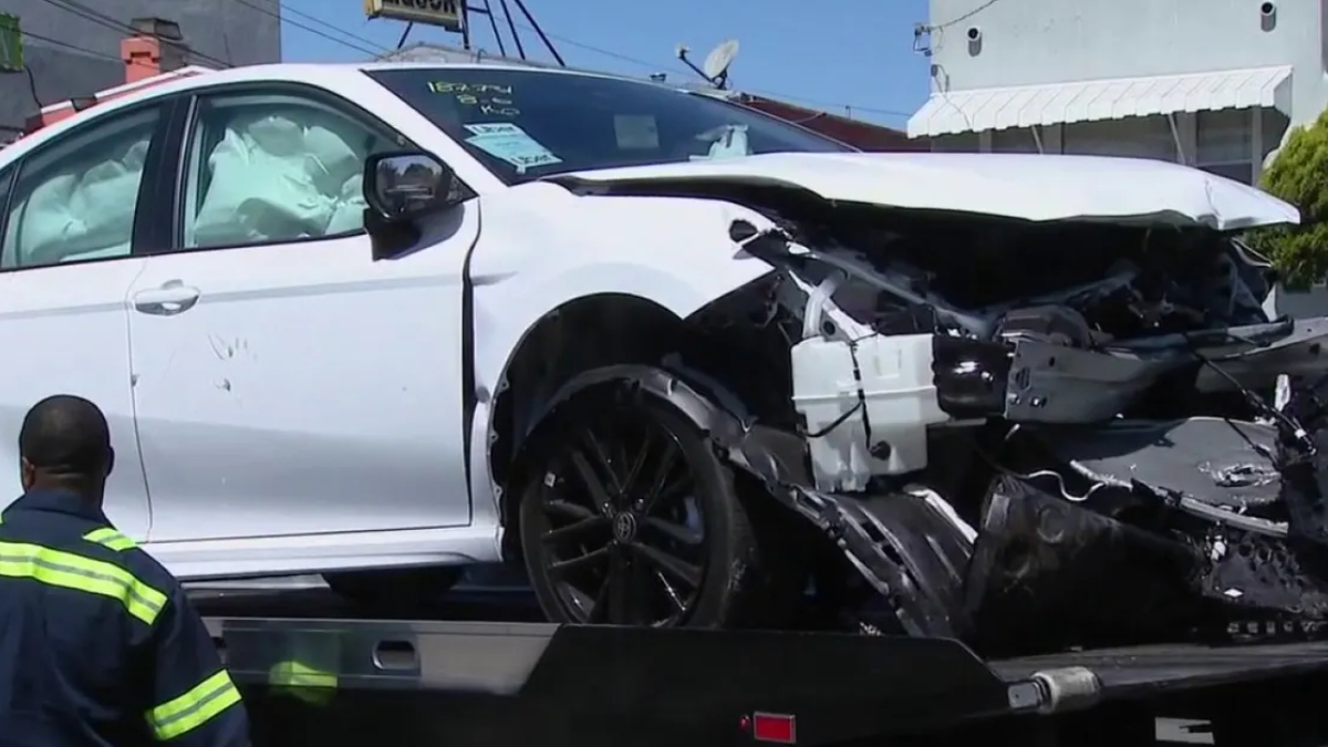 White sedan with severe front-end damage and deployed airbags on a tow truck bed, emergency responder nearby in a neon jacket.