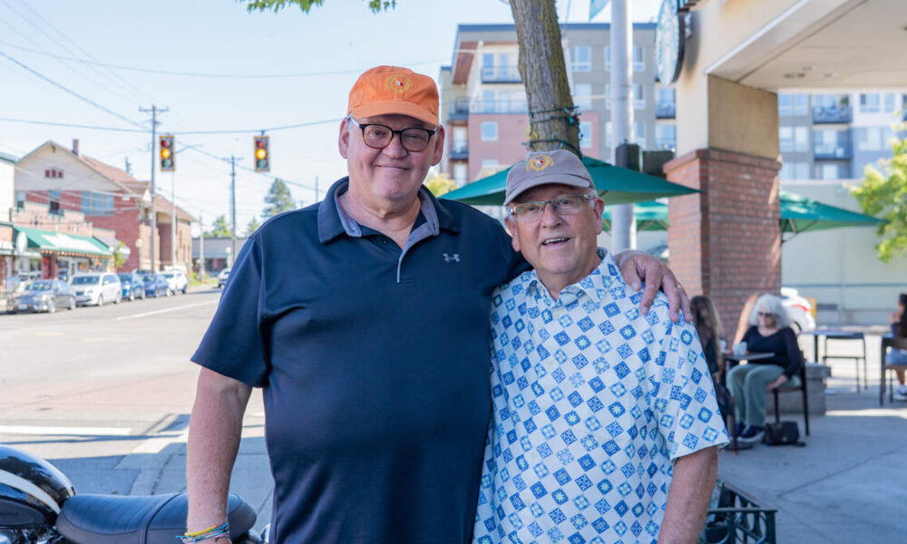 Two smiling men with arms around each other on a sunny city street, cafe seating visible in the background.