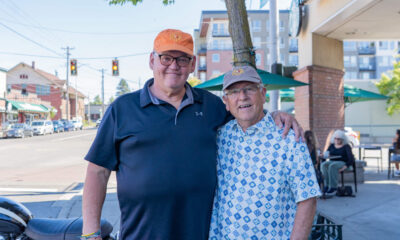 Two smiling men with arms around each other on a sunny city street, cafe seating visible in the background.