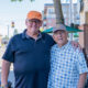 Two smiling men with arms around each other on a sunny city street, cafe seating visible in the background.