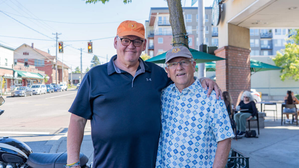 Two smiling men with arms around each other on a sunny city street, cafe seating visible in the background.