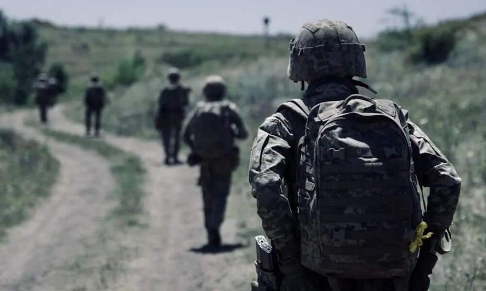Soldier in camouflage gear walking along a dirt path with teammates in the distance.