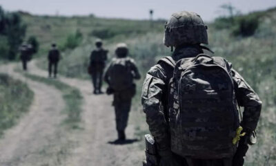 Soldier in camouflage gear walking along a dirt path with teammates in the distance.