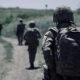 Soldier in camouflage gear walking along a dirt path with teammates in the distance.