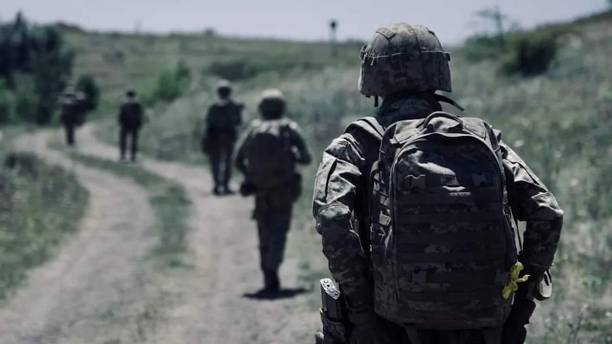 Soldier in camouflage gear walking along a dirt path with teammates in the distance.