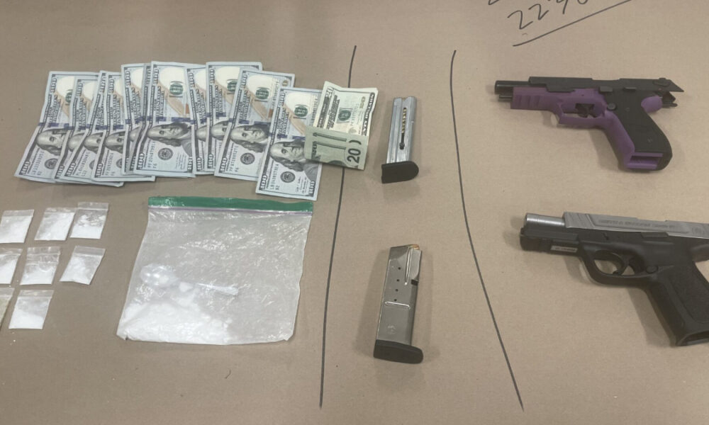 Assorted illegal drugs in small bags, a pile of cash, and two handguns on a brown surface (drug paraphernalia in view).