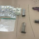 Assorted illegal drugs in small bags, a pile of cash, and two handguns on a brown surface (drug paraphernalia in view).
