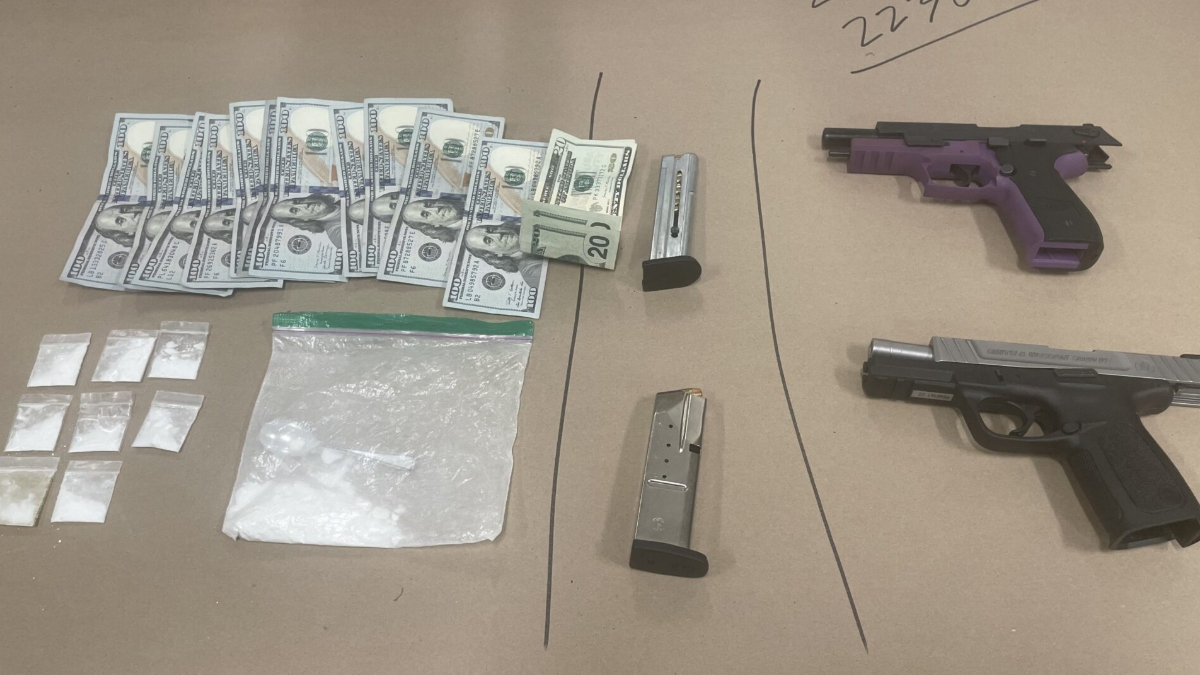 Assorted illegal drugs in small bags, a pile of cash, and two handguns on a brown surface (drug paraphernalia in view).