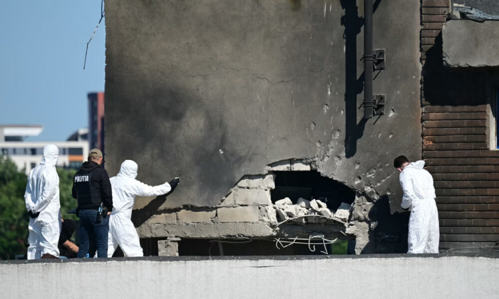 Forensic investigators in white protective suits and a police officer examine a damaged building with a large cracked wall and rubble on a rooftop edge.