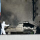 Forensic investigators in white protective suits and a police officer examine a damaged building with a large cracked wall and rubble on a rooftop edge.