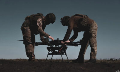 Two soldiers in combat gear inspect a quadcopter drone on the ground outdoors on a field.