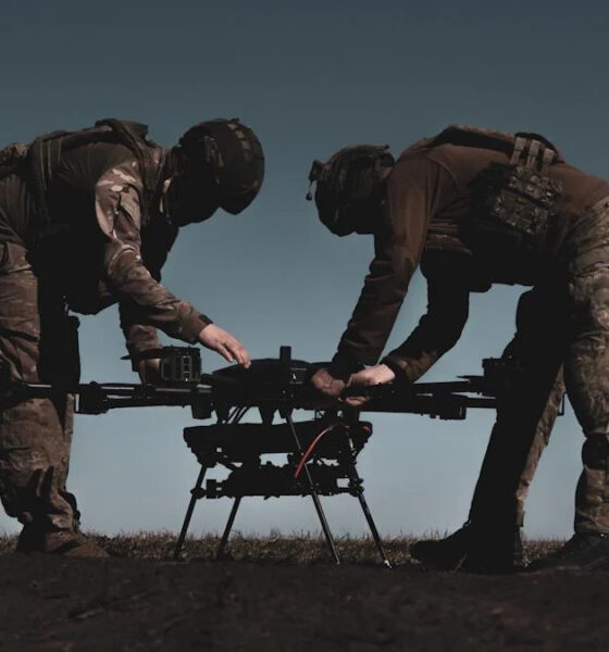 Two soldiers in combat gear inspect a quadcopter drone on the ground outdoors on a field.