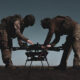Two soldiers in combat gear inspect a quadcopter drone on the ground outdoors on a field.