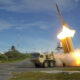 Missile being launched from a mobile launcher on a test range, with bright exhaust plume and smoke at the moment of liftoff.