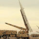 Camouflaged military trucks in a desert with large missiles mounted on launch rails, ready for launch.