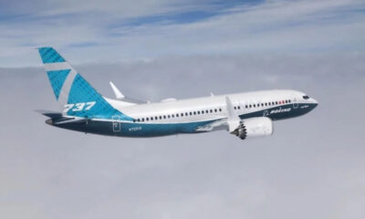 Boeing 737 MAX in flight with blue-turquoise tail and white fuselage, engine visible under the wing.