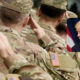 Soldiers in camouflage saluting; inset shows a blond-haired man in a suit with flags in the background.