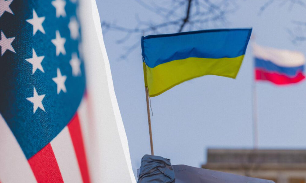 Close-up of the US flag on the left and a Ukrainian flag on a pole, with a blurred Russian flag in the background.