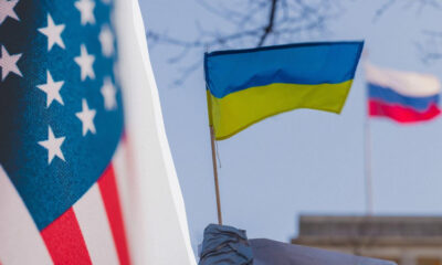 Close-up of the US flag on the left and a Ukrainian flag on a pole, with a blurred Russian flag in the background.