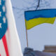Close-up of the US flag on the left and a Ukrainian flag on a pole, with a blurred Russian flag in the background.