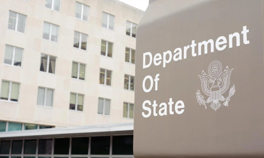 Close-up of a gray Department of State sign with the U.S. seal, set against a beige multi-story building in the background.