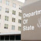 Close-up of a gray Department of State sign with the U.S. seal, set against a beige multi-story building in the background.