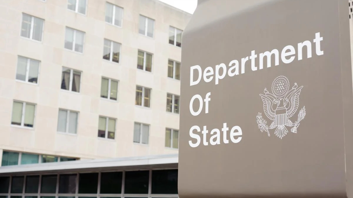 Close-up of a gray Department of State sign with the U.S. seal, set against a beige multi-story building in the background.