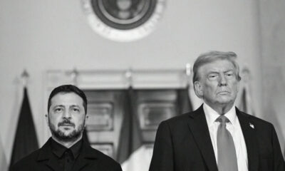 Two men in dark suits stand side by side in a formal government room with flags and an official seal above.