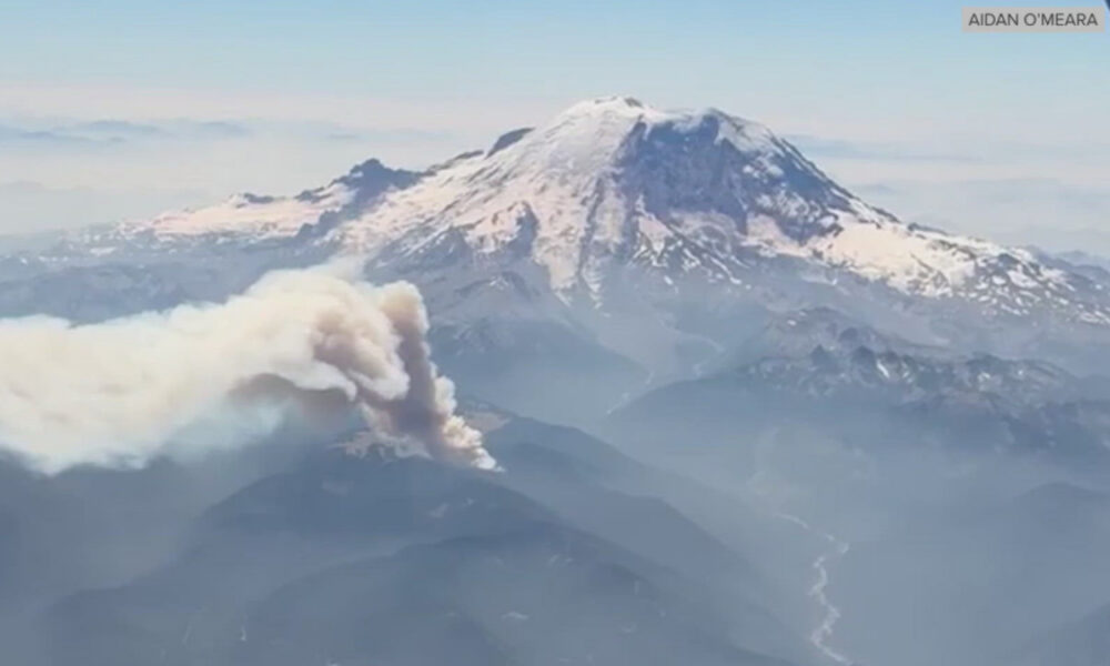 Aerial view of a snow‑capped mountain with a thick ash plume rising from its slopes, indicating a volcanic eruption.