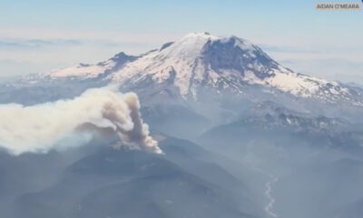 Aerial view of a snow‑capped mountain with a thick ash plume rising from its slopes, indicating a volcanic eruption.
