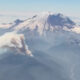 Aerial view of a snow‑capped mountain with a thick ash plume rising from its slopes, indicating a volcanic eruption.