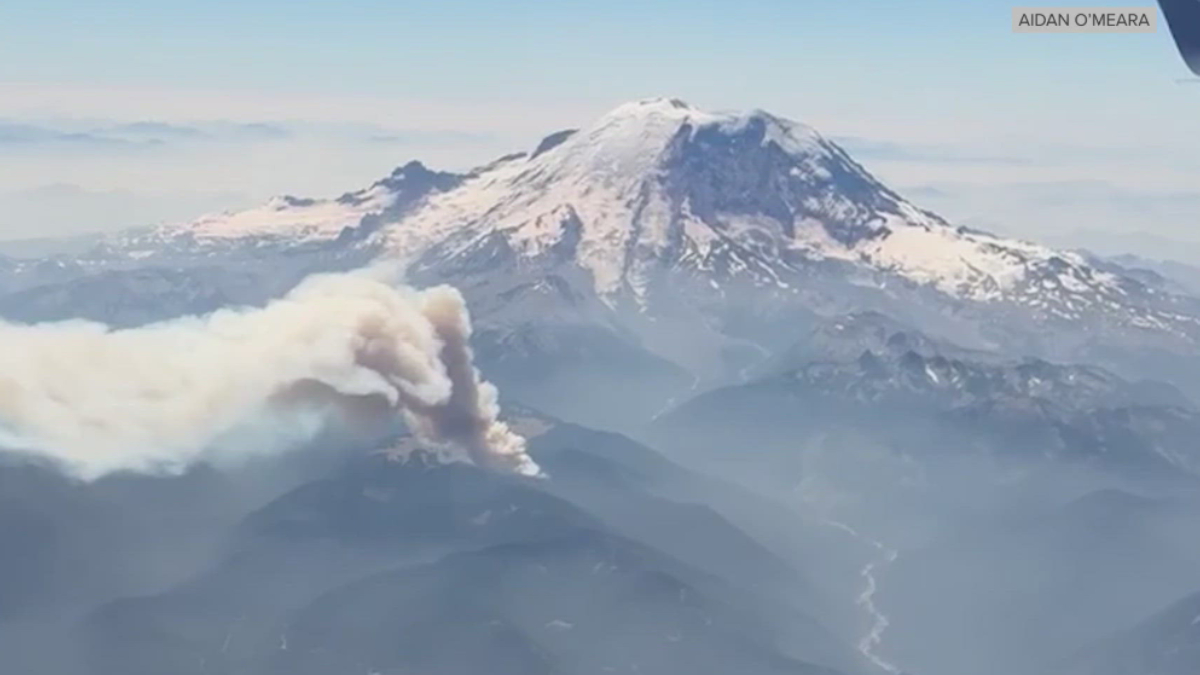 Aerial view of a snow‑capped mountain with a thick ash plume rising from its slopes, indicating a volcanic eruption.