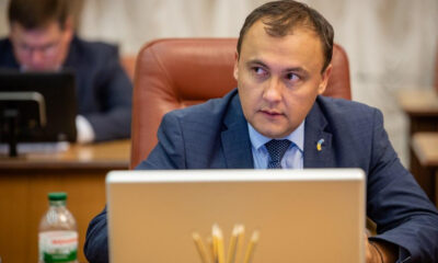 Man in a blue suit sits at a desk with a laptop, looking to the side during a formal meeting.