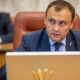 Man in a blue suit sits at a desk with a laptop, looking to the side during a formal meeting.
