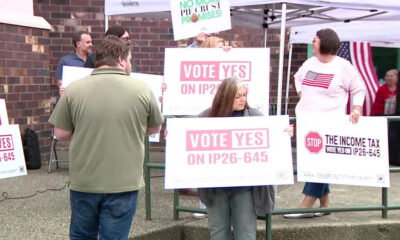 Group of people at an outdoor rally holding signs that say 'Vote Yes on IP26-645' in pink letters, near a tent and brick wall roadside.