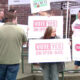 Group of people at an outdoor rally holding signs that say 'Vote Yes on IP26-645' in pink letters, near a tent and brick wall roadside.