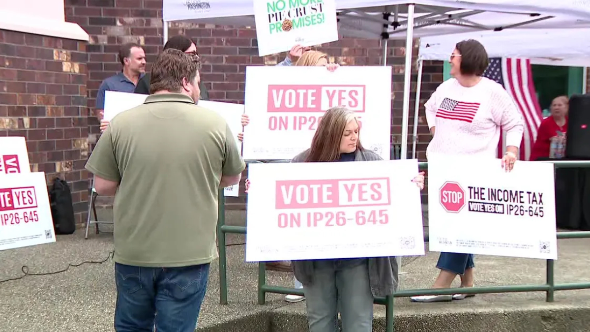 Group of people at an outdoor rally holding signs that say 'Vote Yes on IP26-645' in pink letters, near a tent and brick wall roadside.