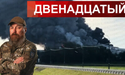 Bearded Ukrainian soldier in camouflage with arms crossed stands in front of a large smoke plume rising from a fire near a road, with a red Cyrillic banner above.