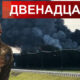 Bearded Ukrainian soldier in camouflage with arms crossed stands in front of a large smoke plume rising from a fire near a road, with a red Cyrillic banner above.