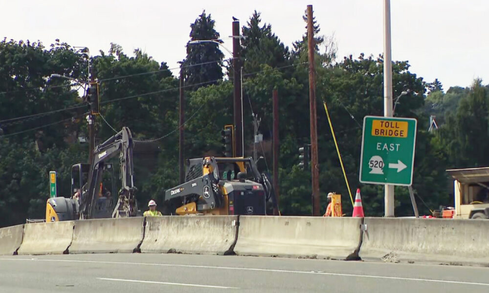 Highway construction zone with a concrete barrier, two excavators, a worker in a yellow helmet, and a green Toll Bridge East sign (Route 520).