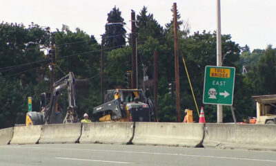 Highway construction zone with a concrete barrier, two excavators, a worker in a yellow helmet, and a green Toll Bridge East sign (Route 520).