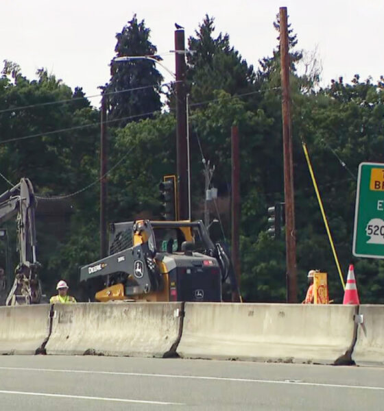 Highway construction zone with a concrete barrier, two excavators, a worker in a yellow helmet, and a green Toll Bridge East sign (Route 520).