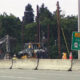 Highway construction zone with a concrete barrier, two excavators, a worker in a yellow helmet, and a green Toll Bridge East sign (Route 520).