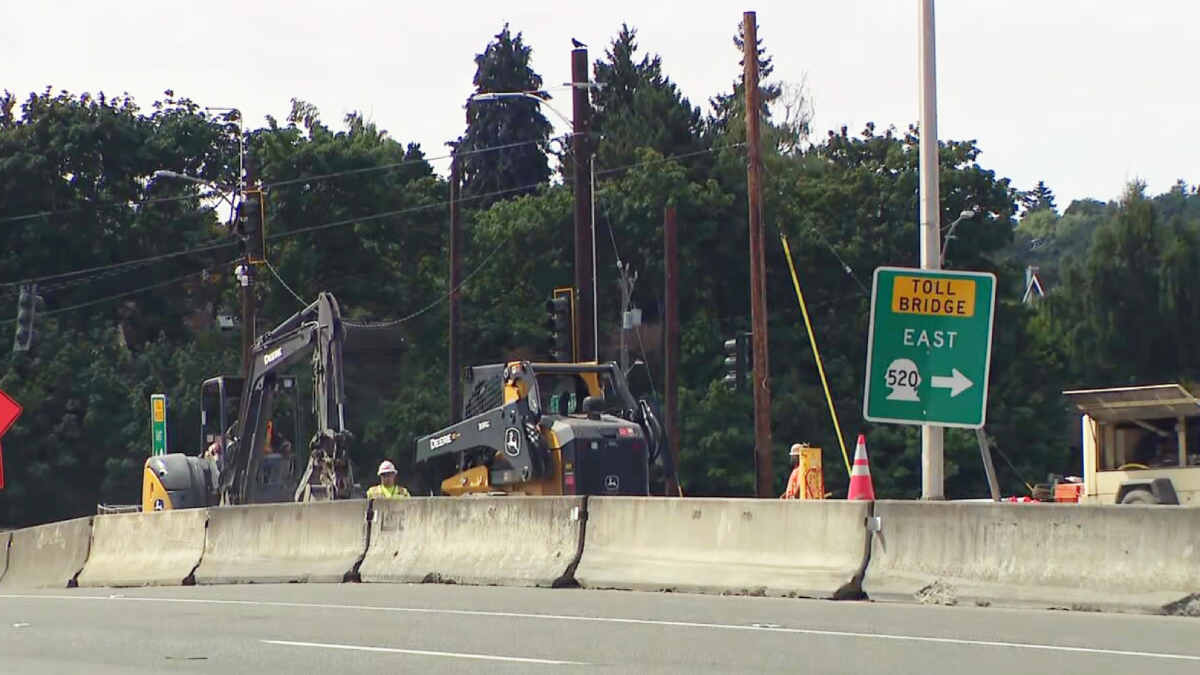 Highway construction zone with a concrete barrier, two excavators, a worker in a yellow helmet, and a green Toll Bridge East sign (Route 520).