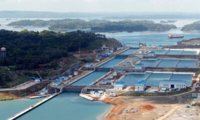 Large lock system with lined concrete basins and blue-roofed buildings beside a river, ships visible in the distance.