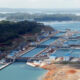 Large lock system with lined concrete basins and blue-roofed buildings beside a river, ships visible in the distance.