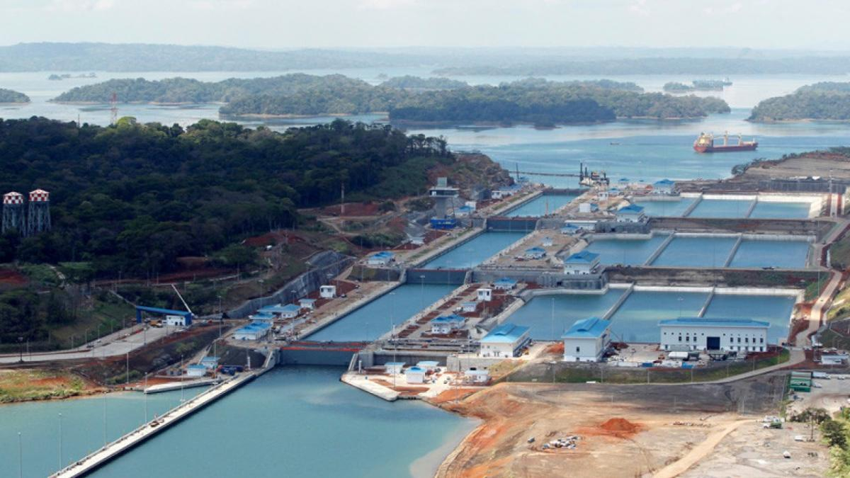 Large lock system with lined concrete basins and blue-roofed buildings beside a river, ships visible in the distance.