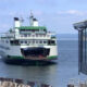 White and green passenger ferry approaching a pier, with the rear loading ramp open and vehicles visible on deck